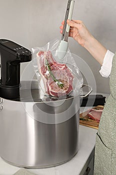 Woman putting vacuum packed meat into pot with sous vide cooker in kitchen, closeup. Thermal immersion circulator