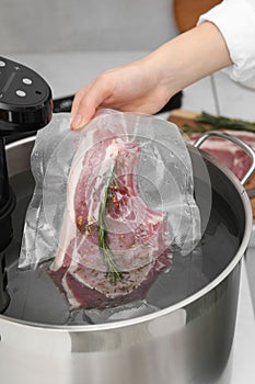 Woman putting vacuum packed meat into pot with sous vide cooker in kitchen, closeup. Thermal immersion circulator