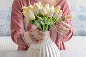 Woman putting white tulips flowers in vase sitting at the living room coffee table. Composing bouquet. Lifestyle.