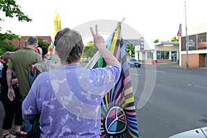 Woman with rainbow flag in crowd gives peace sign to passing motorists