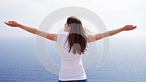 Woman raise arms up at cliff against blue sea. Female traveleer outstretching hands