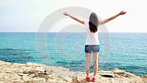 Woman raise arms up at cliff against blue sea. Female traveleer outstretching hands in slow motion