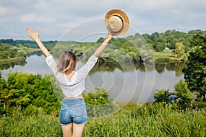 Woman raising arms feeling free and happy walking by summer lake. Girl admires natural landscape view