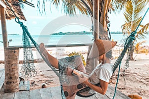 Woman reading book on hammock tropical beach, real people getting away from it all, traditional south asian hat, palm trees, teal