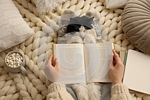 Woman reading book and holding adorable cat on knitted blanket, top view