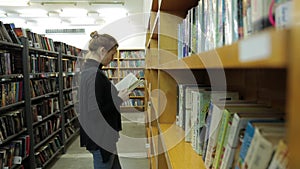 Woman is reading a book in library. She is in the corridor between the books.