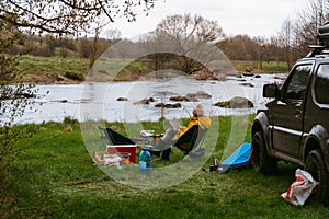 A woman is reading a book. An off-road vehicle with a roof rack. camping equipment. View on river. Concept of vacation and travel