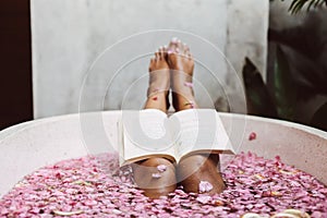 Woman reading book while relaxing in bath tub with flower petals