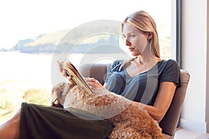 Woman Reading Book Relaxing In Chair By Window At Home With Pet Dog