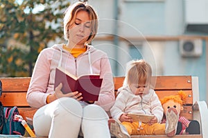 Woman is reading a book sitting on a bench. A child is sitting nearby and watching cartoons on a smartphone. The concept