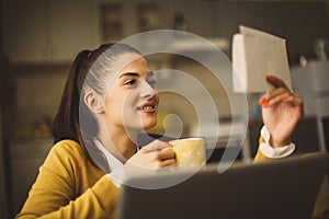 Woman reading document using laptop and drinking coffee at