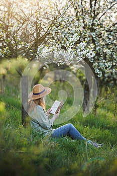 Woman is reading in her notebook diary and relaxing under blooming cherry tree