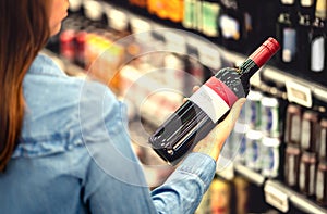 Woman reading the label of red wine bottle in liquor store or alcohol section of supermarket. Shelf full of alcoholic beverages.