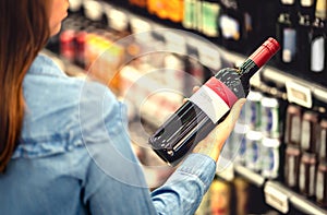 Woman reading the label of red wine bottle in liquor store or alcohol section of supermarket. Shelf full of alcoholic beverages. photo