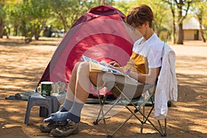 Woman reading magazine while relaxing in camping site. Tent, chairs and camping gears. Outdoor activities in summer. Adventure tra