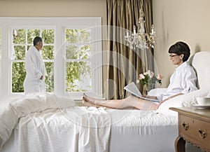 Woman Reading Newspaper And Man Standing Near Window In Bedroom