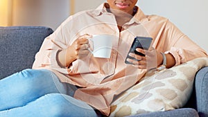 Woman reading a phone with funny memes on social media, drinking coffee and relaxing on sofa. Closeup of happy black