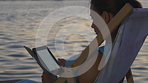 Woman reads an e-book at sunset near glaring water