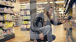 Woman reads the label on the package and bottle at the supermarket. Looking for products from the low shelves and put it