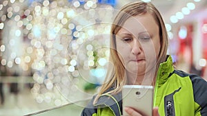 A woman reads a smartphone message and compresses lips. She stands in a mall, amid a light bulb out of focus. Christmas