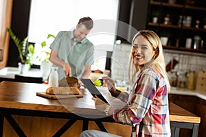 A woman reads from a tablet at the kitchen counter while a man holding an tomatoes smiles at her