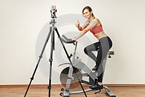 Woman recording tutorial video on cell phone on tripod, riding exercise bike and showing thumb up.