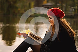 Woman in a red beret reading book on wooden pontoon. Autumn season.