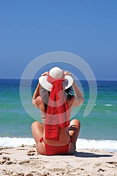 Woman in red bikini sitting on beach adjusting hat