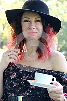 woman with red curls of hair, dressed in a black dress and a black hat, with a Cup of tea in her hand, vertical