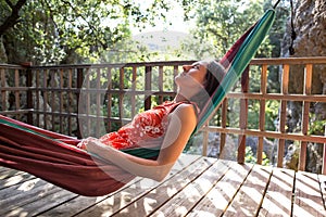 A woman in a red dress lies in a hammock on the terrace of a wooden house and enjoys the view