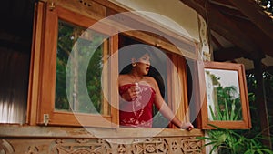 Woman in red dress looking out from a rustic window, with greenery in the background