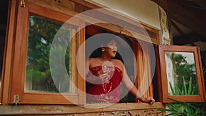 Woman in red dress looking out from a rustic window, with greenery in the background