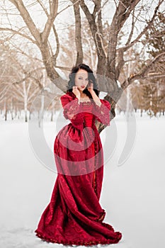 woman in a red dress of the Rococo era stands in the snow against the background of a winter forest