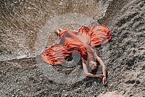 Woman red dress sea. Female dancer in a long red dress posing on a beach with rocks on sunny day