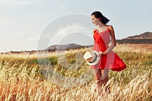 Woman in a red dress walking on the field on a warm summer evening. Yellow grass at sunset, the girl holding a hat in his hands