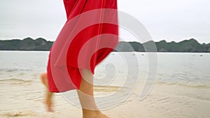The woman in a red dress walks on the coast of the island. Rear view. Ocean surf. Tropical island.Traces of legs on sand