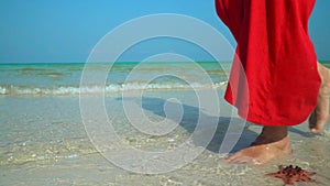 The woman in a red dress walks on the coast with white sand. Woman in red dress on the tropical beach. Starfish on phu