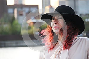 Woman with red hair and a classic black hat by the city river