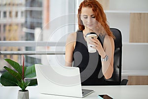 Woman with red hair is drinking coffee and talking on the phone at the workplace in the office