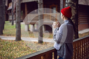 woman in a red hat and scarf and a mug stands at a wooden house in the woods