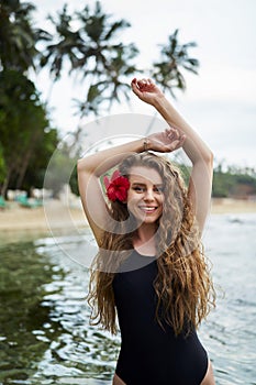 Woman with red hibiscus in hair enjoys tropical sea. Female in black swimsuit at beach escape. Solo traveler explores