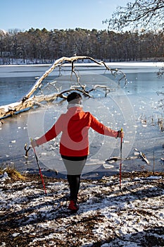 A woman in a red jacket practices Nordic walking in the woods and along the lake