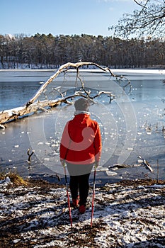 A woman in a red jacket practices Nordic walking in the woods and along the lake