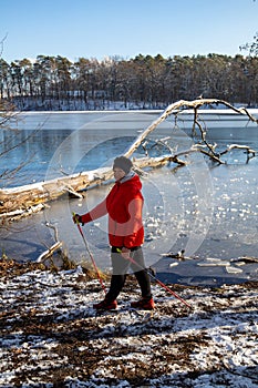 A woman in a red jacket practices Nordic walking in the woods and along the lake