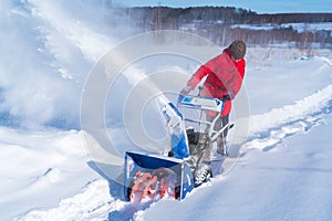 A woman in a red jacket removes snow from a rural road with a snowblower in winter after a snowfall