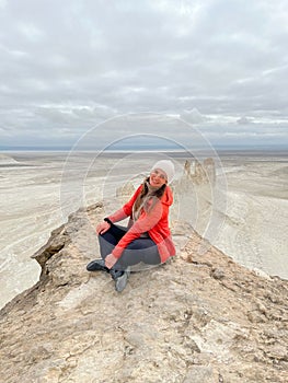 A woman in red jacket sits on a rocky peak overlooking the expansive, arid Bozzhyra landscape