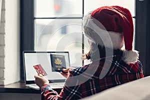Woman in a red santa claus hat holding credit card and american passport using laptop for making order sitting near window.