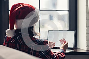 Woman in a red santa claus hat holding credit card using laptop for making order sitting near window. Christmas