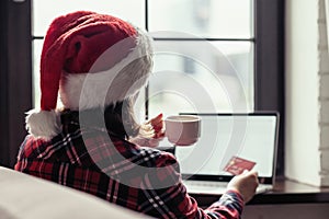 Woman in a red santa claus hat holding credit card using laptop for making order sitting near window. Christmas