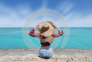 A woman in a red shirt and jeans posing with a big hat with a blue sea view